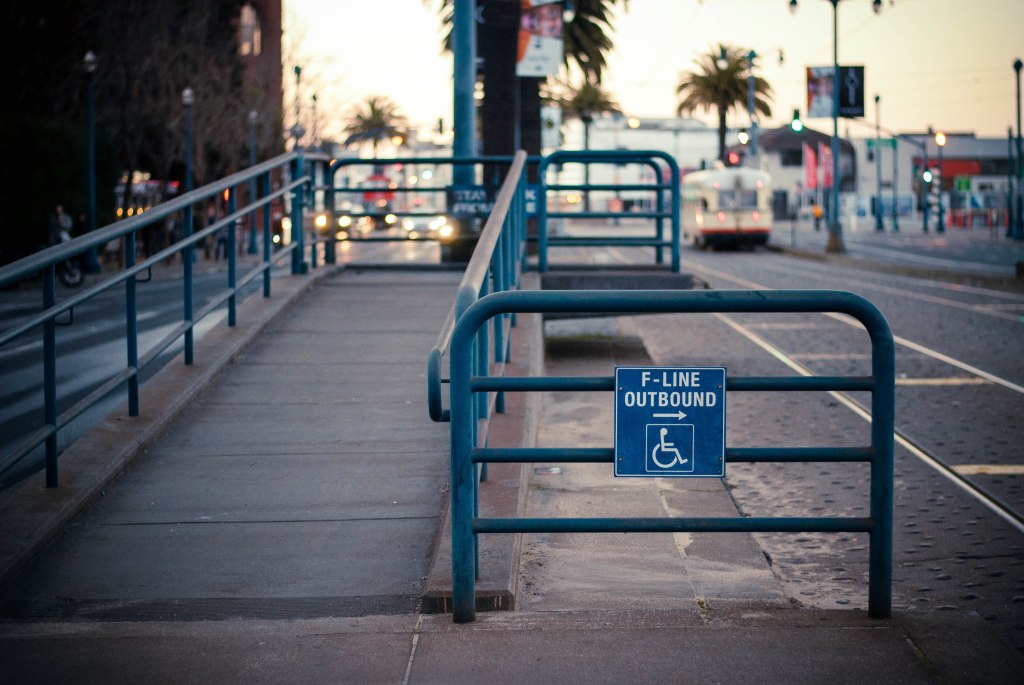 Photo (by Rafael Castillo) of an outdoor San Francisco Muni light rail stop on Market Street, with a wheelchair-accessible ramp. A sign on the railing says "F-line outbound" and has the standard wheelchair symbol.